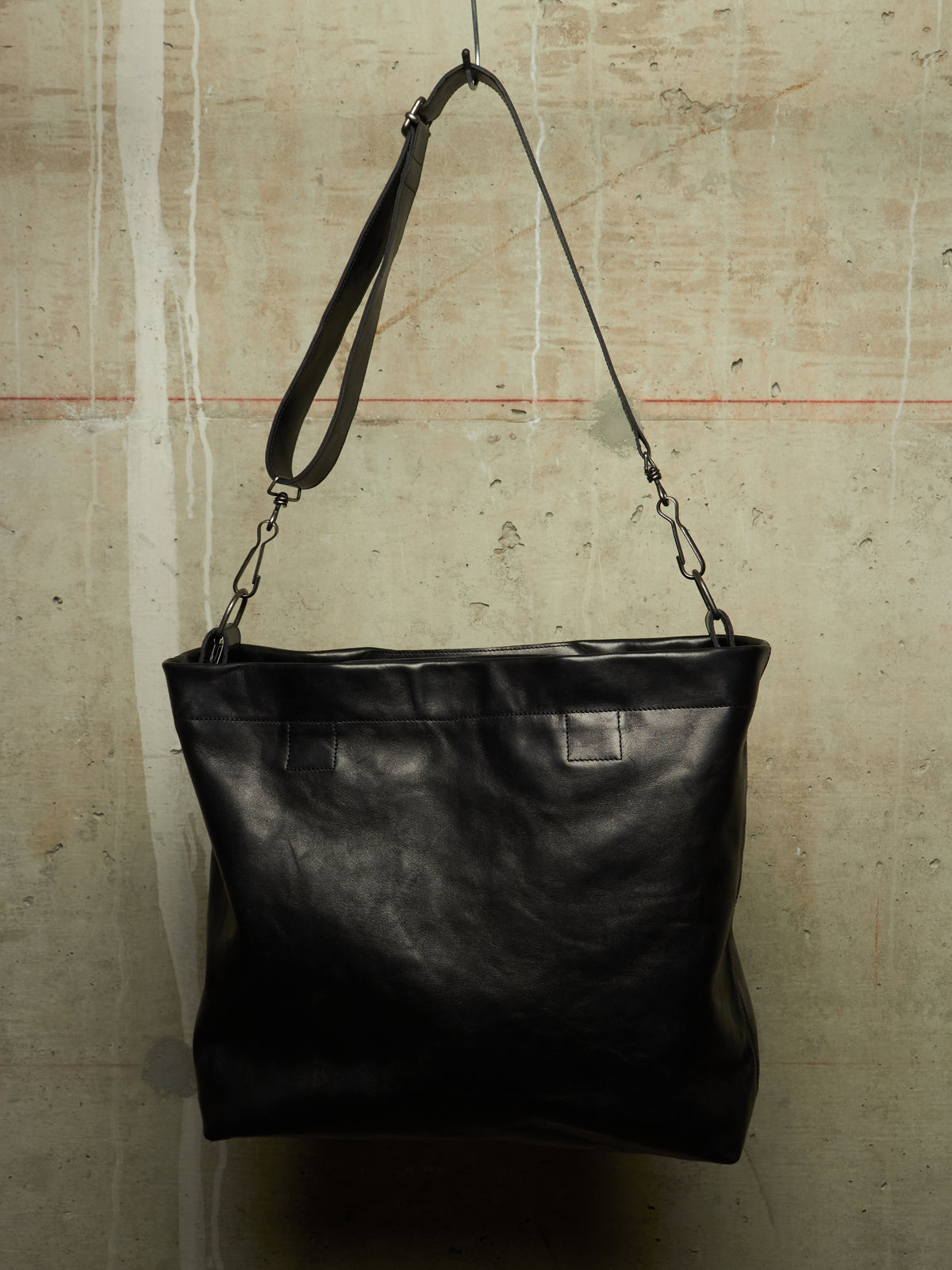 A black leather shoulder bag with a wide top opening and a long strap, hanging against a textured concrete wall.
