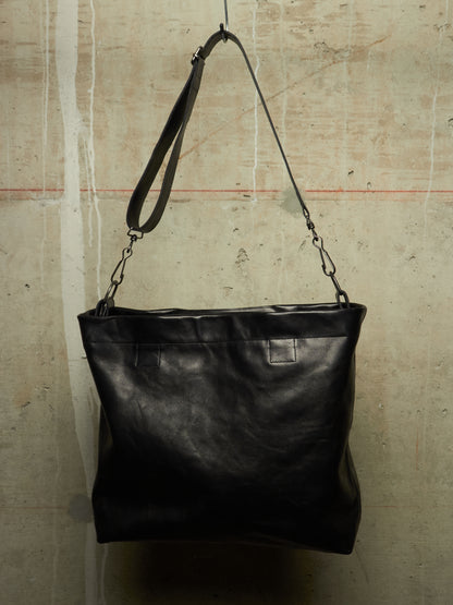 A black leather shoulder bag with a wide top opening and a long strap, hanging against a textured concrete wall.