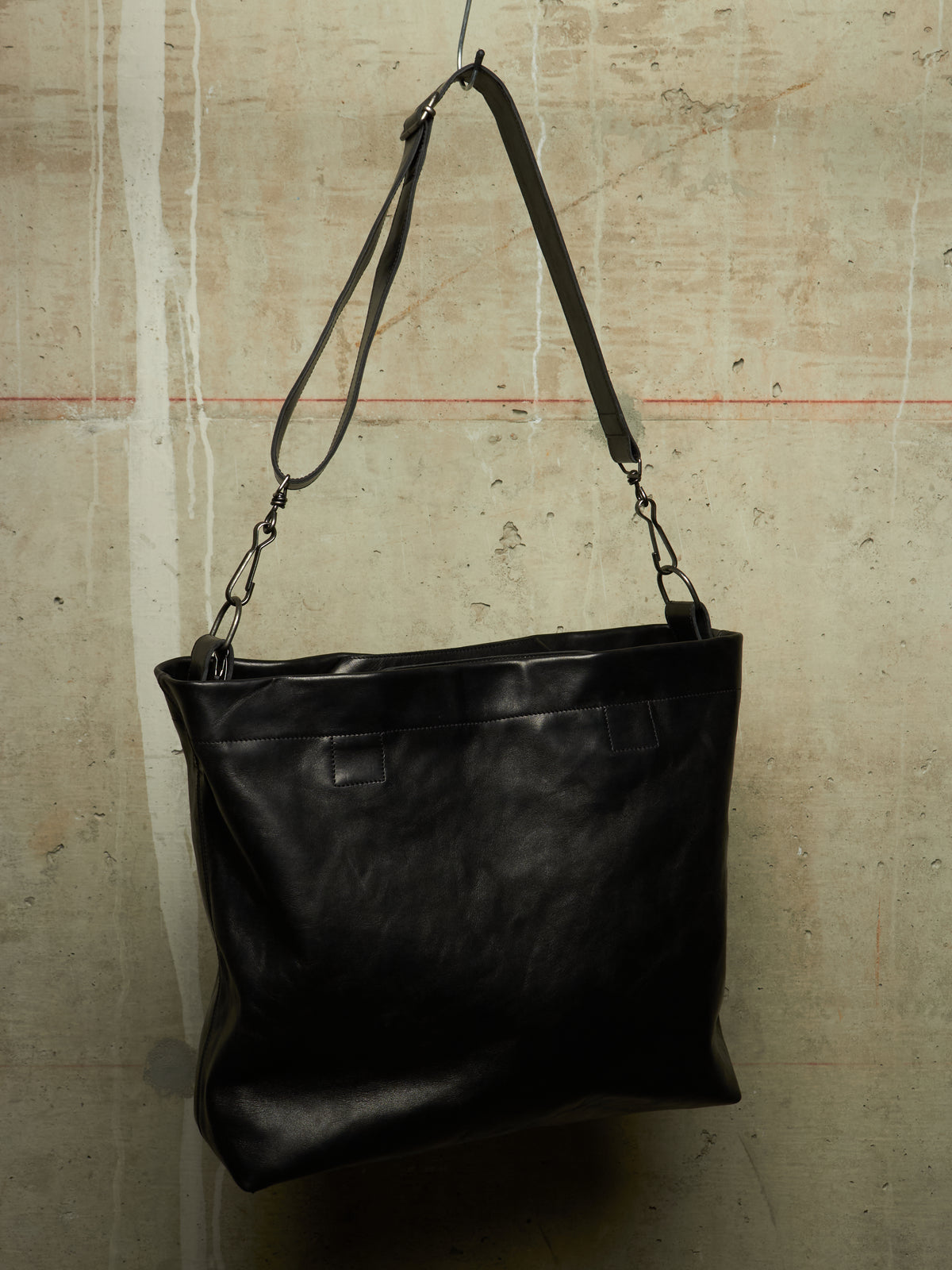 A black leather shoulder bag with a long strap, hanging against a textured concrete wall.
