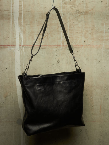 A black leather shoulder bag with a long strap, hanging against a textured concrete wall.