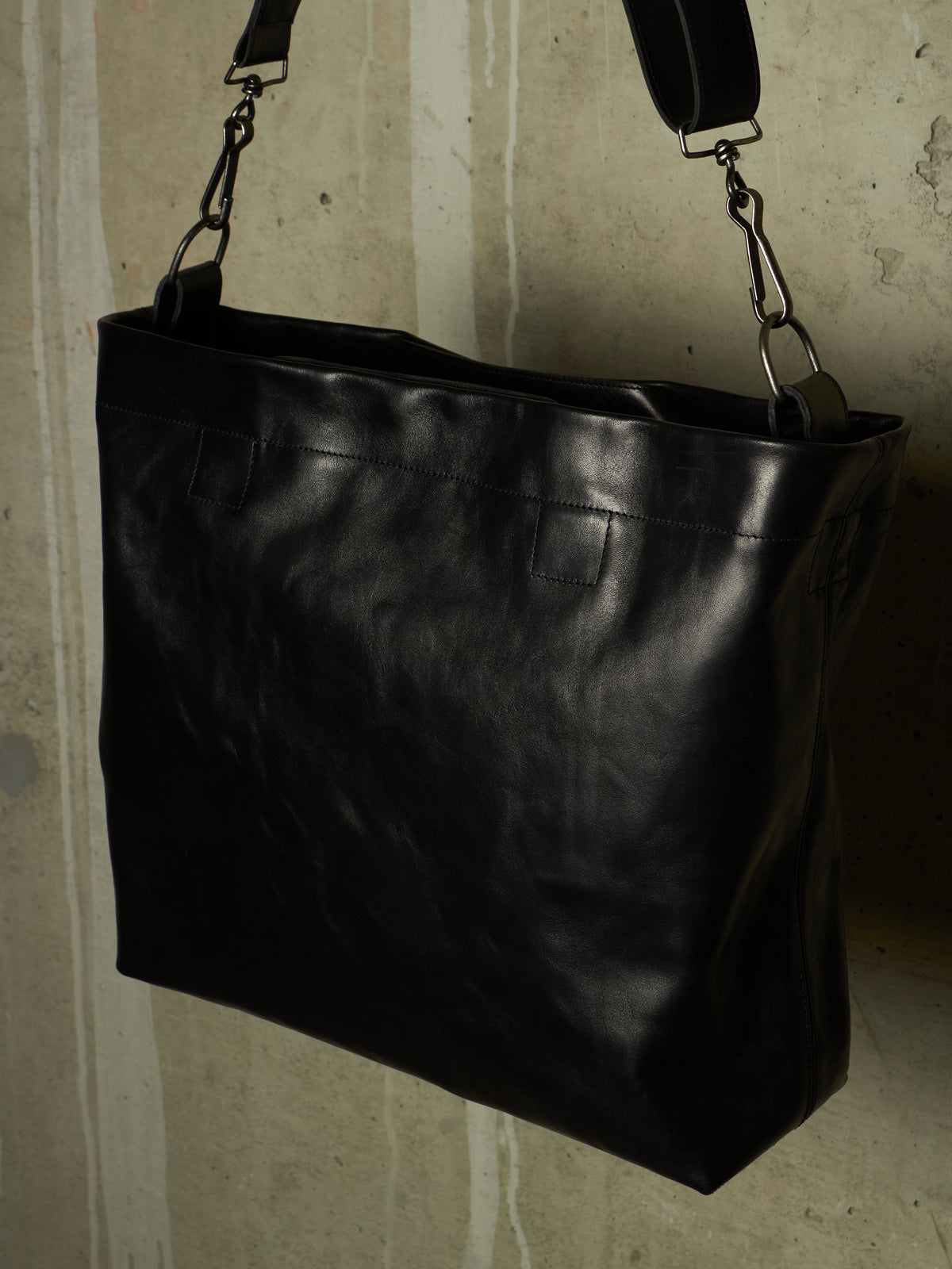 A large black leather shoulder bag with a wide top opening and a long adjustable strap, hanging against a textured concrete wall.