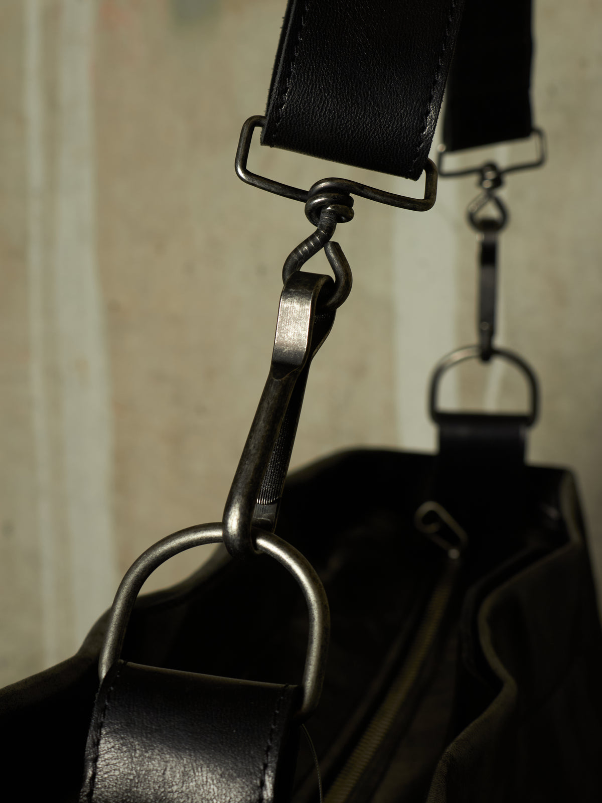 Close-up detail of a black leather bag with dark metal hardware, showing a strap attachment and D-ring on a neutral background.