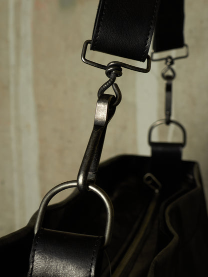 Close-up detail of a black leather bag with dark metal hardware, showing a strap attachment and D-ring on a neutral background.