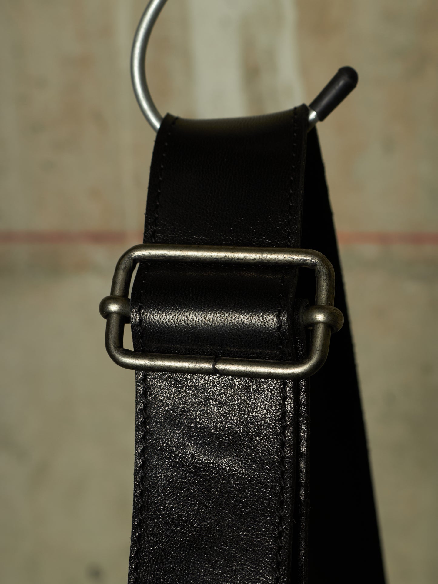 A close-up of a black leather belt with a silver-toned metal buckle and a black plastic keeper loop, hanging against a textured beige background.
