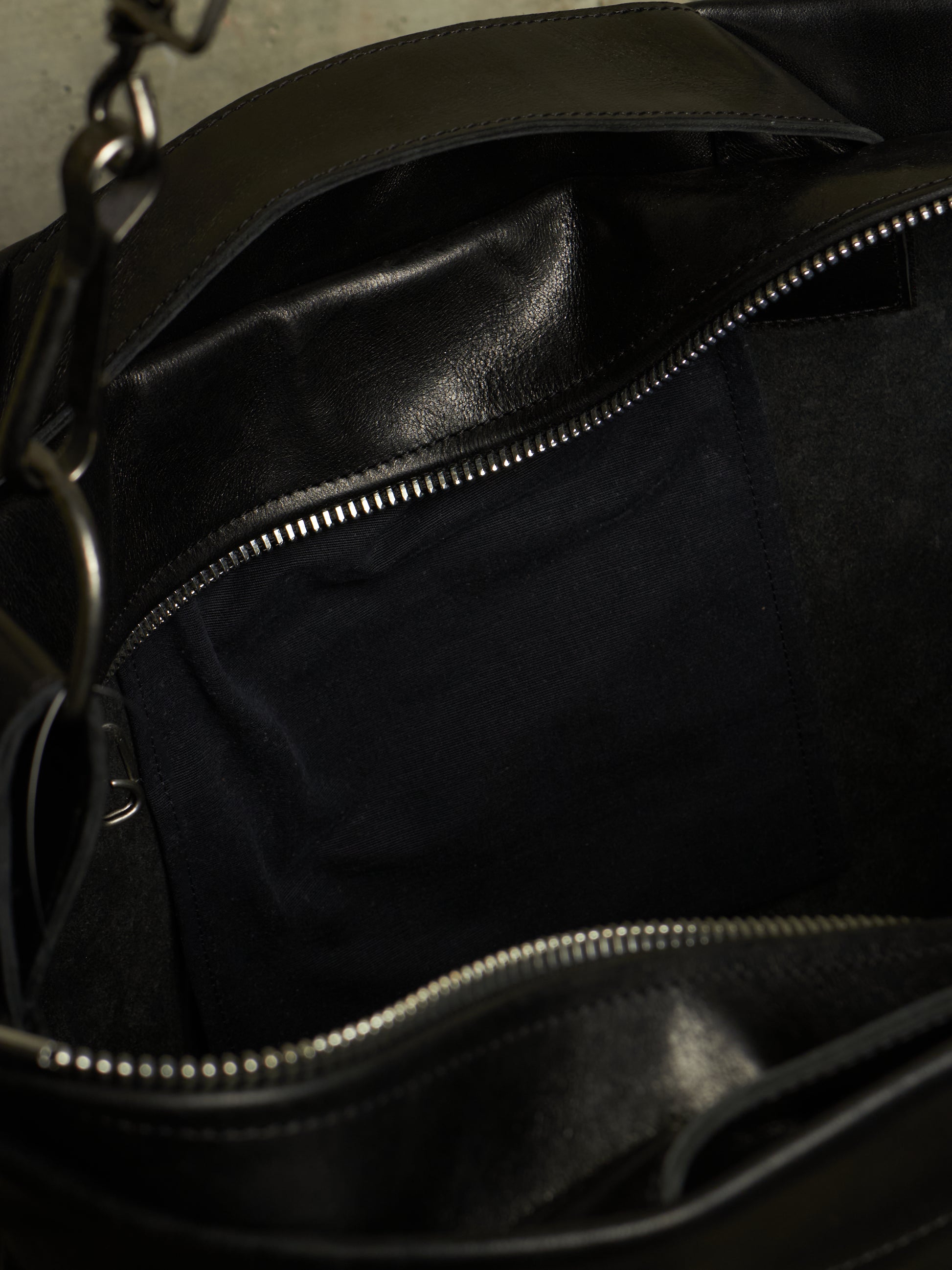Close-up of the interior of a black leather bag, showing a dark fabric lining, silver zipper tracks, and part of a metal chain strap.