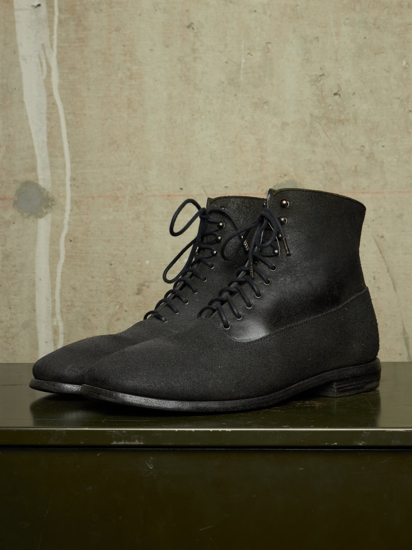 A pair of black lace-up ankle boots with a matte textured finish, displayed on a dark green surface against a textured beige wall.