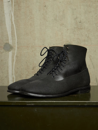 A pair of black lace-up ankle boots with a matte textured finish, displayed on a dark green surface against a textured beige wall.