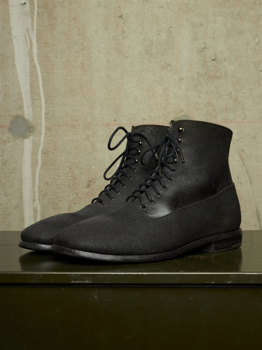 A pair of black lace-up ankle boots with a matte textured finish, displayed on a dark green surface against a textured beige wall.