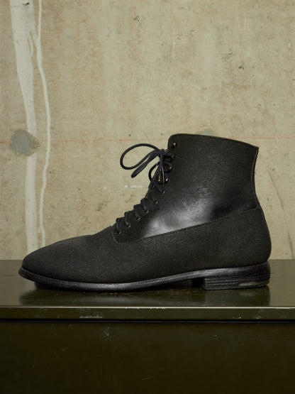 A black lace-up ankle boot with a matte suede-like finish, displayed in profile on a dark green surface against a textured beige wall.