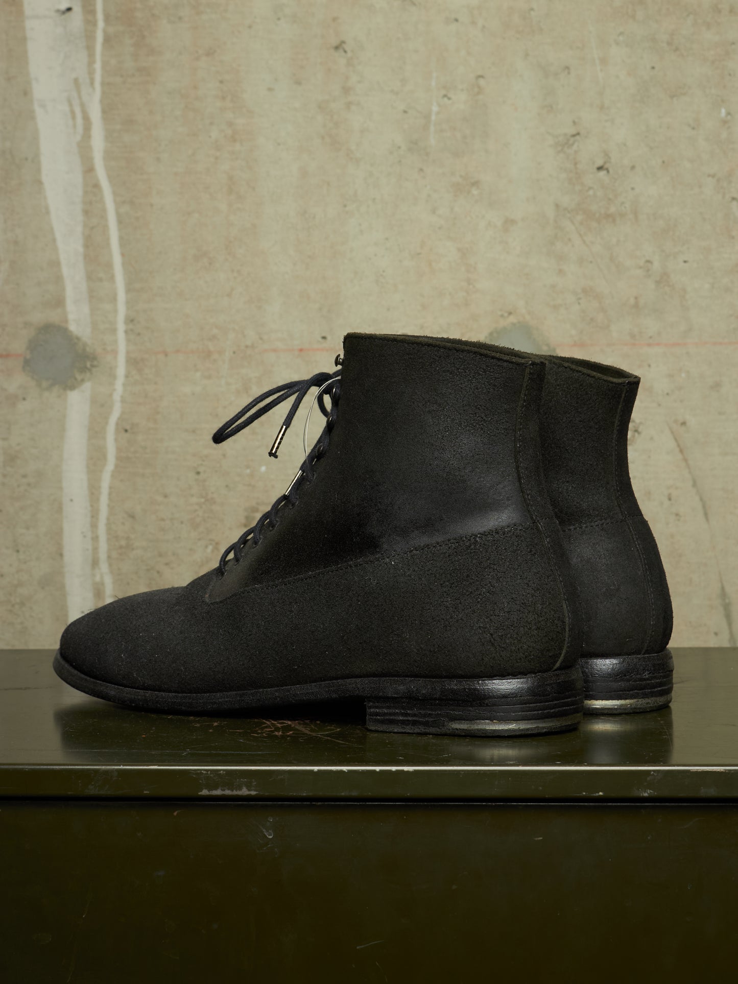 A pair of black suede lace-up ankle boots displayed on a dark green surface against a textured beige concrete wall.