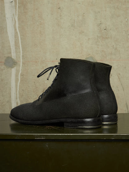 A pair of black suede lace-up ankle boots displayed on a dark green surface against a textured beige concrete wall.