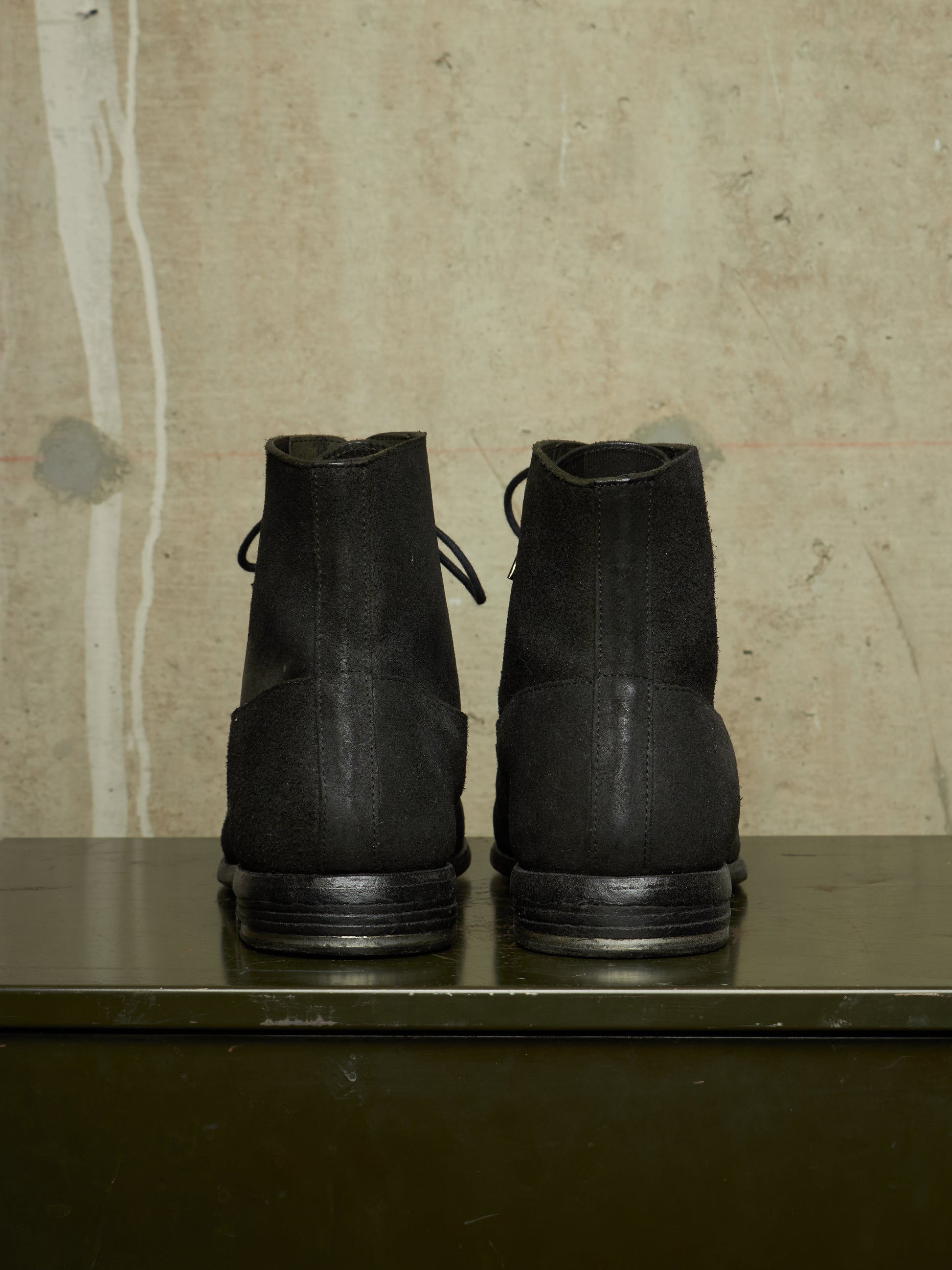 A pair of black lace-up ankle boots shown from the back, resting on a dark green metal surface against a textured beige concrete wall.