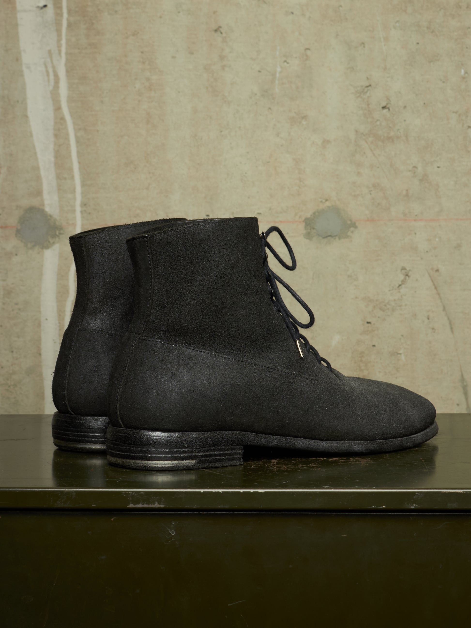 A pair of black suede lace-up ankle boots displayed on a dark green metal surface against a textured beige concrete wall.