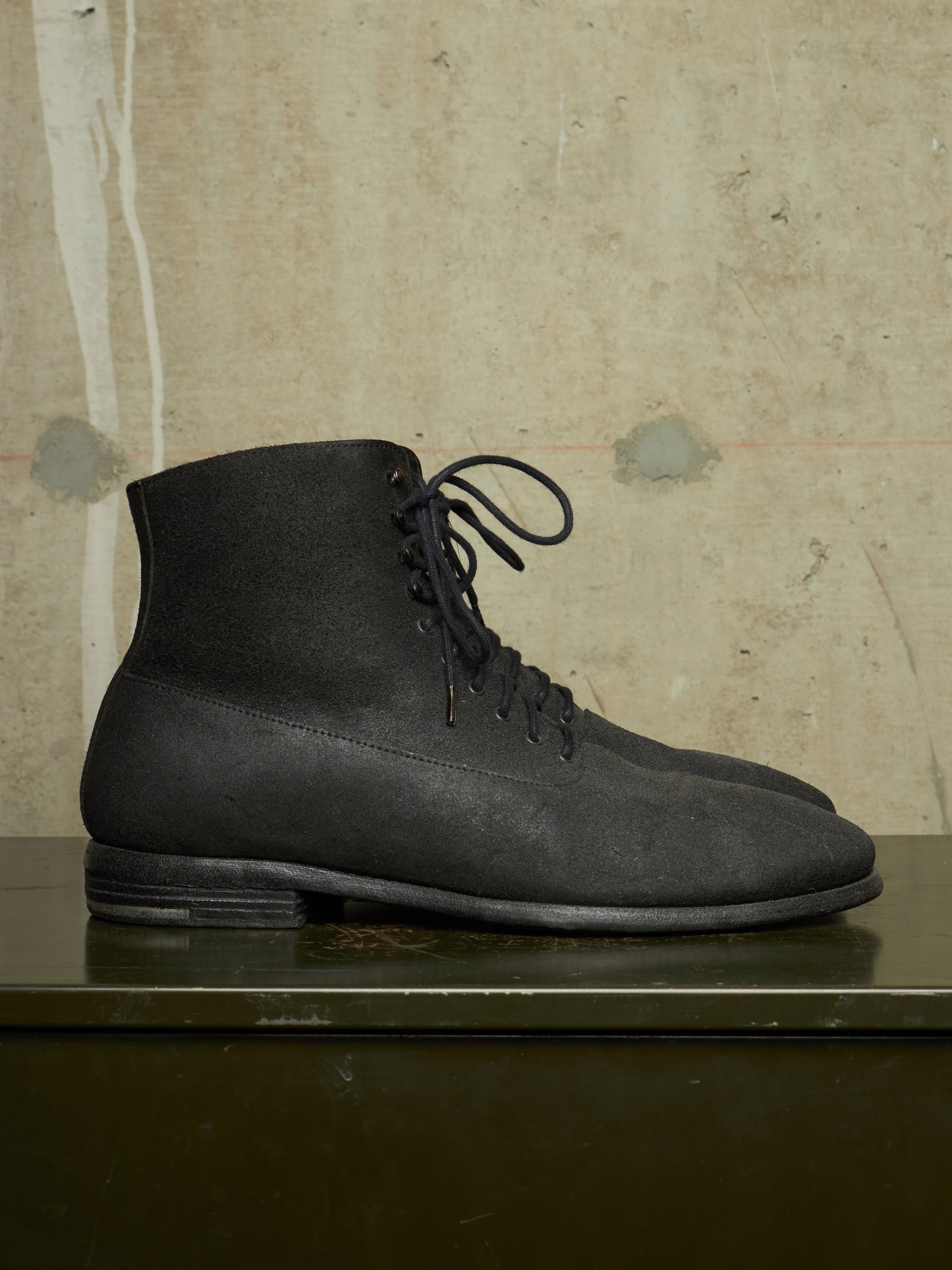 A pair of black suede lace-up ankle boots displayed on a dark green reflective surface against a textured beige concrete wall.