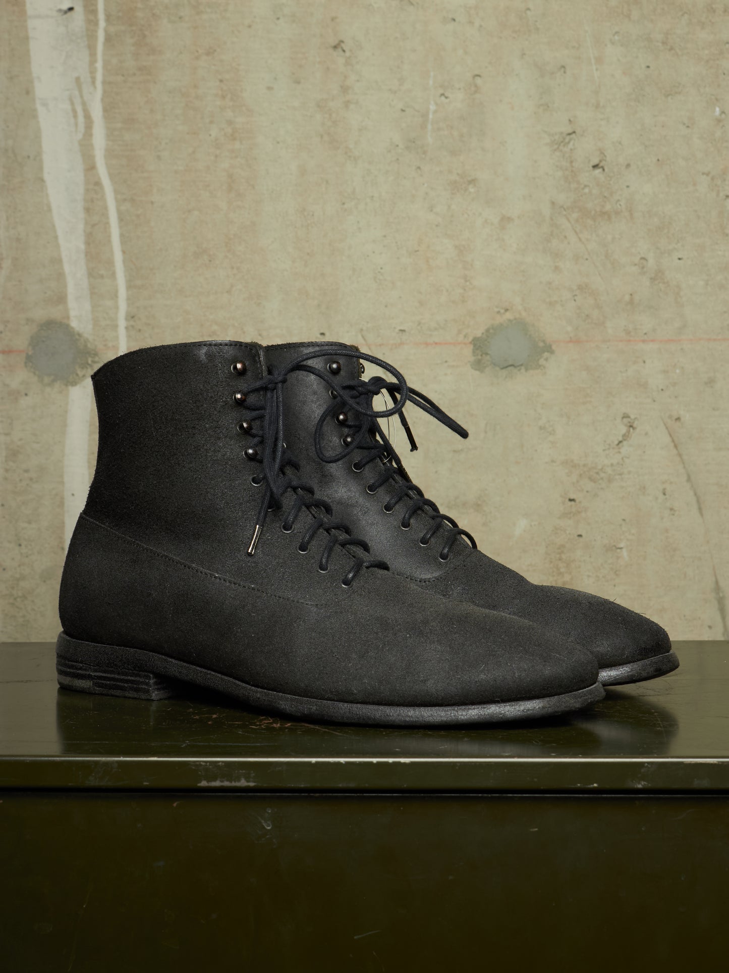 A pair of dark charcoal gray suede lace-up ankle boots with metal eyelets, displayed on a dark green surface against a textured beige concrete wall.