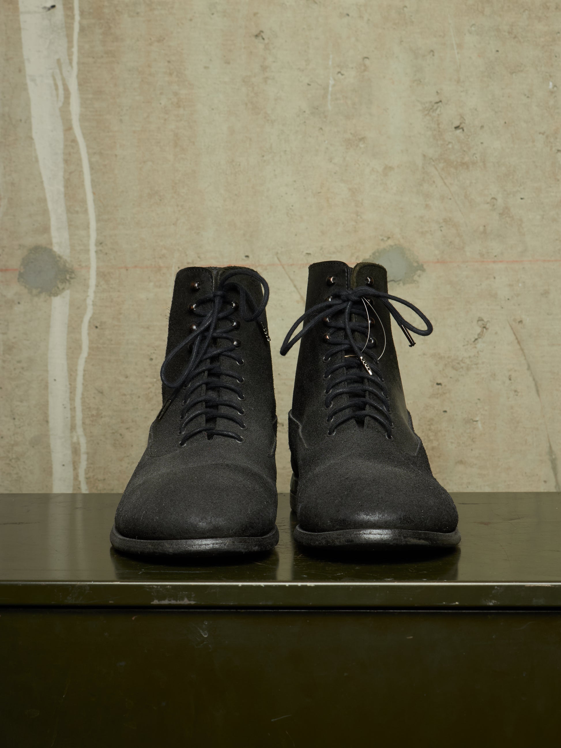 A pair of black lace-up ankle boots with a matte finish, displayed side-by-side on a dark green surface against a textured beige concrete wall.