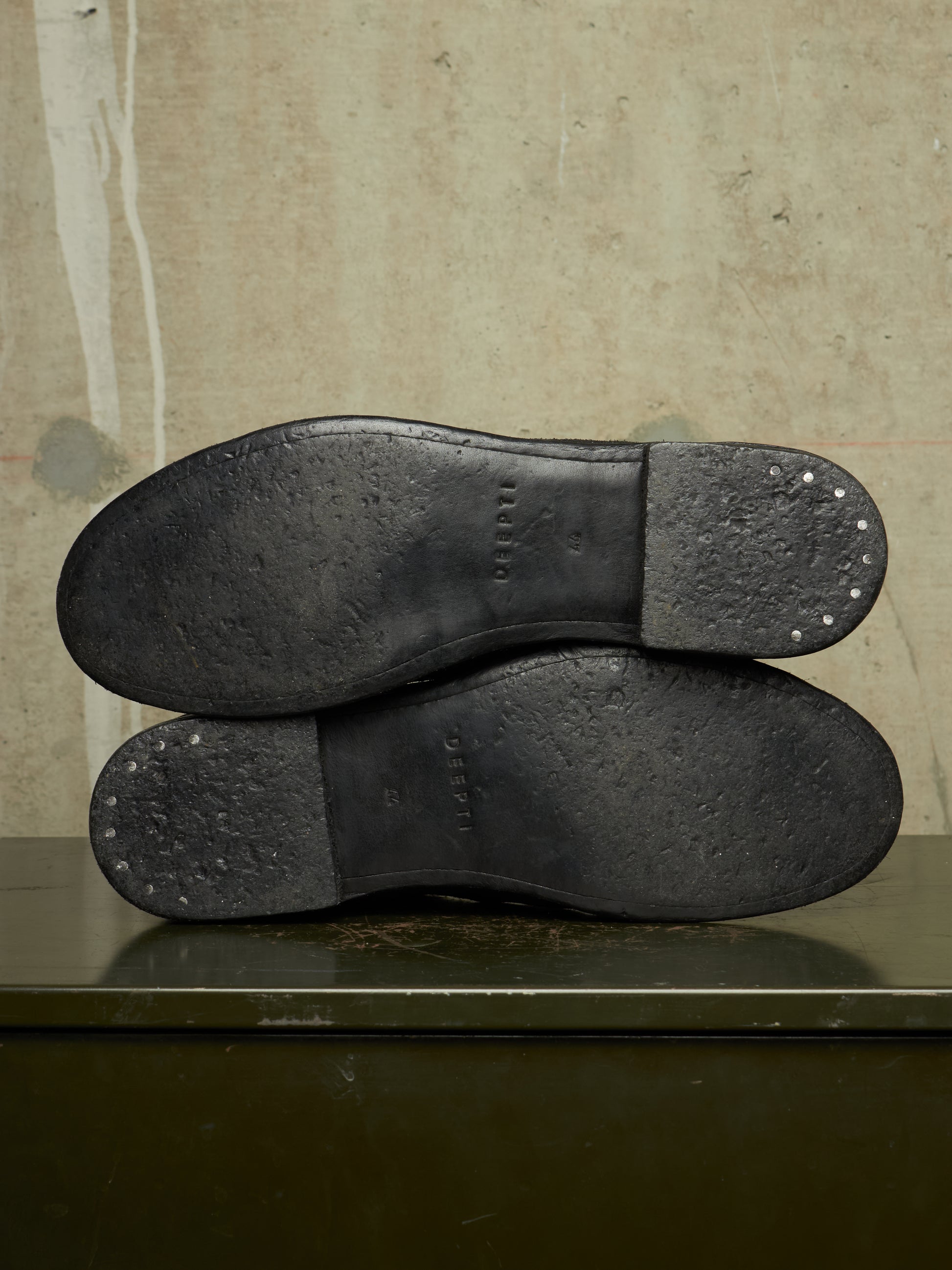 The soles of a pair of black leather shoes, showing the tread and heel caps with small metal studs, resting on a dark green surface against a textured beige wall.