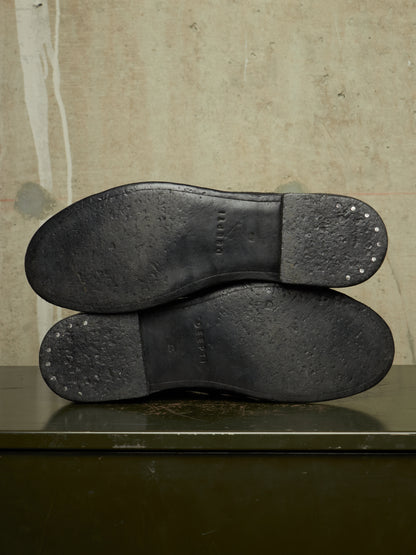 The soles of a pair of black leather shoes, showing the tread and heel caps with small metal studs, resting on a dark green surface against a textured beige wall.