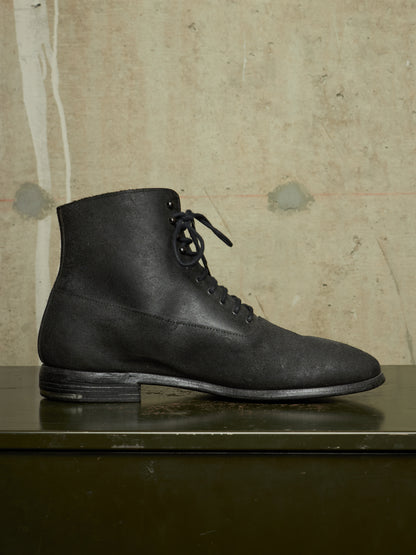 A single black leather lace-up ankle boot with a low block heel, displayed in profile on a dark green reflective surface against a textured beige concrete wall.