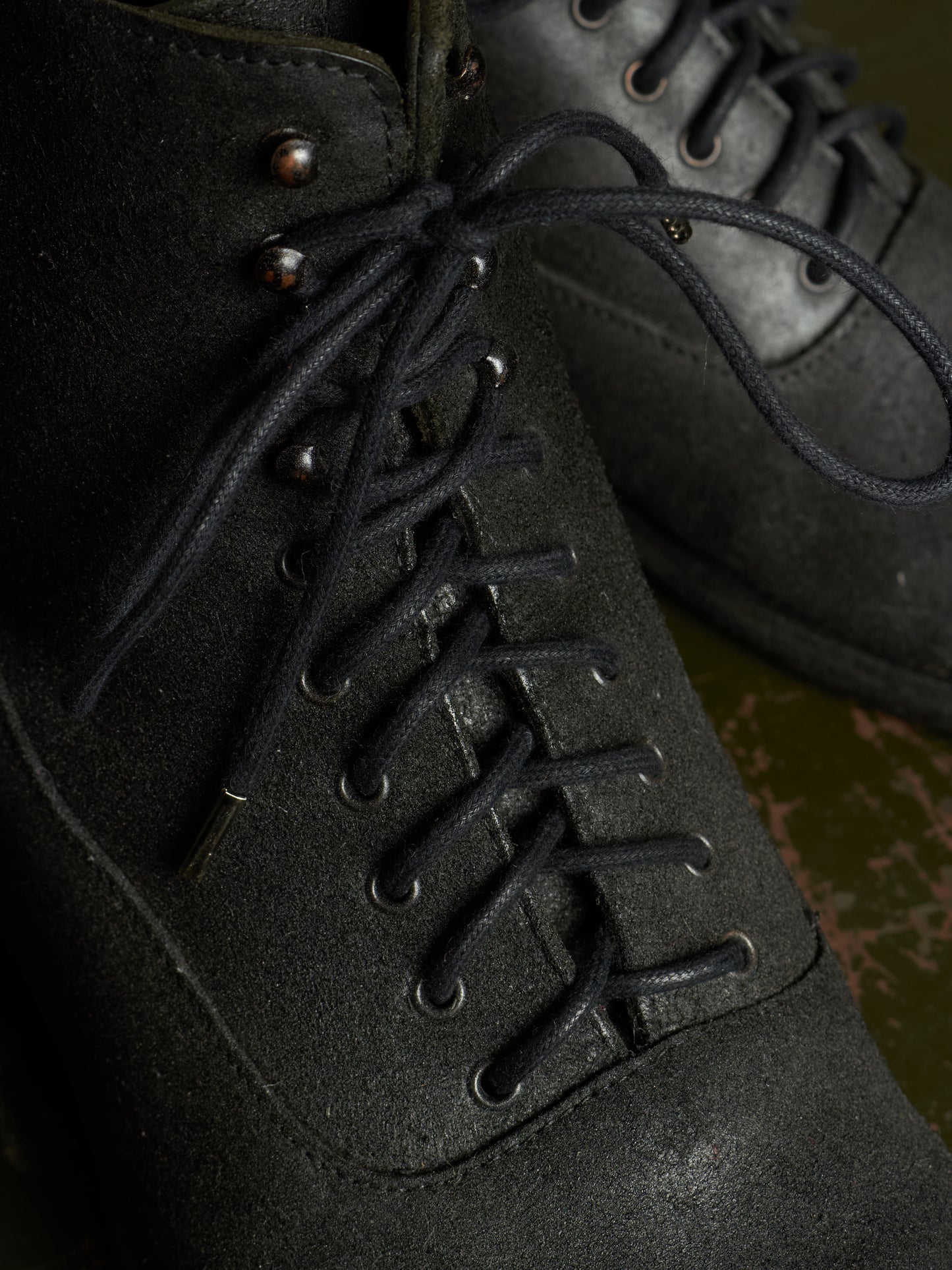 Close-up of a dark charcoal gray suede lace-up boot with black laces and metal eyelets, shown against a dark background.