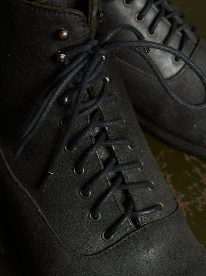 Close-up of a dark charcoal gray suede lace-up boot with black laces and metal eyelets, shown against a dark background.