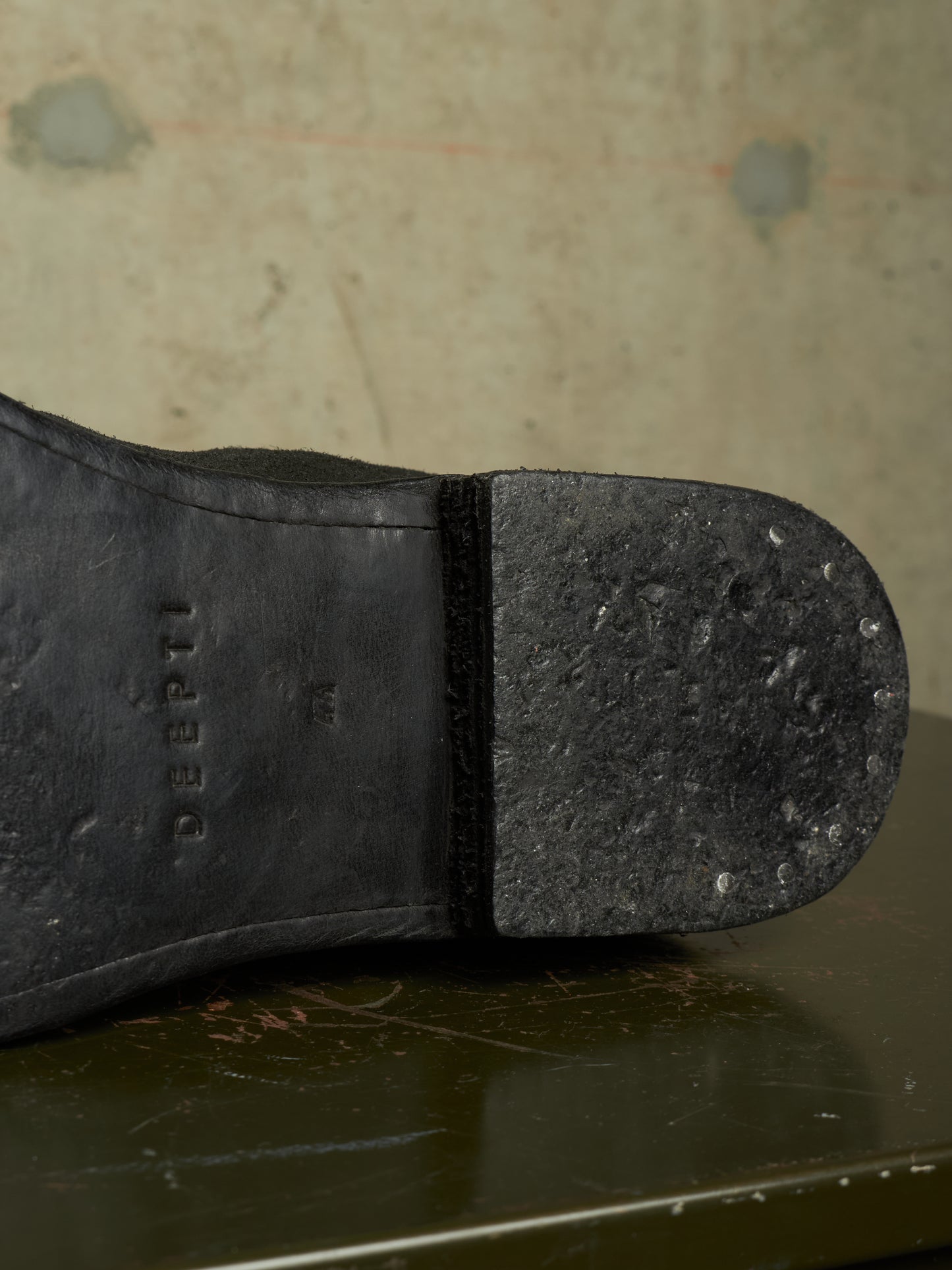 Close-up of the sole of a black leather boot, showing the brand name 'DEEPTI' embossed on the heel and a textured rubber outsole, resting on a dark reflective surface against a textured beige wall.
