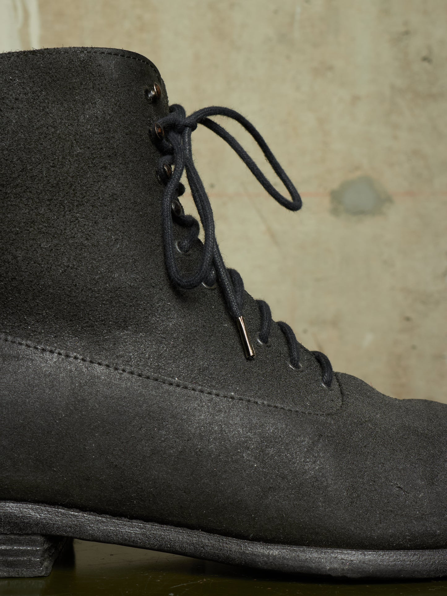 A close-up of a dark gray suede lace-up boot against a textured beige background.