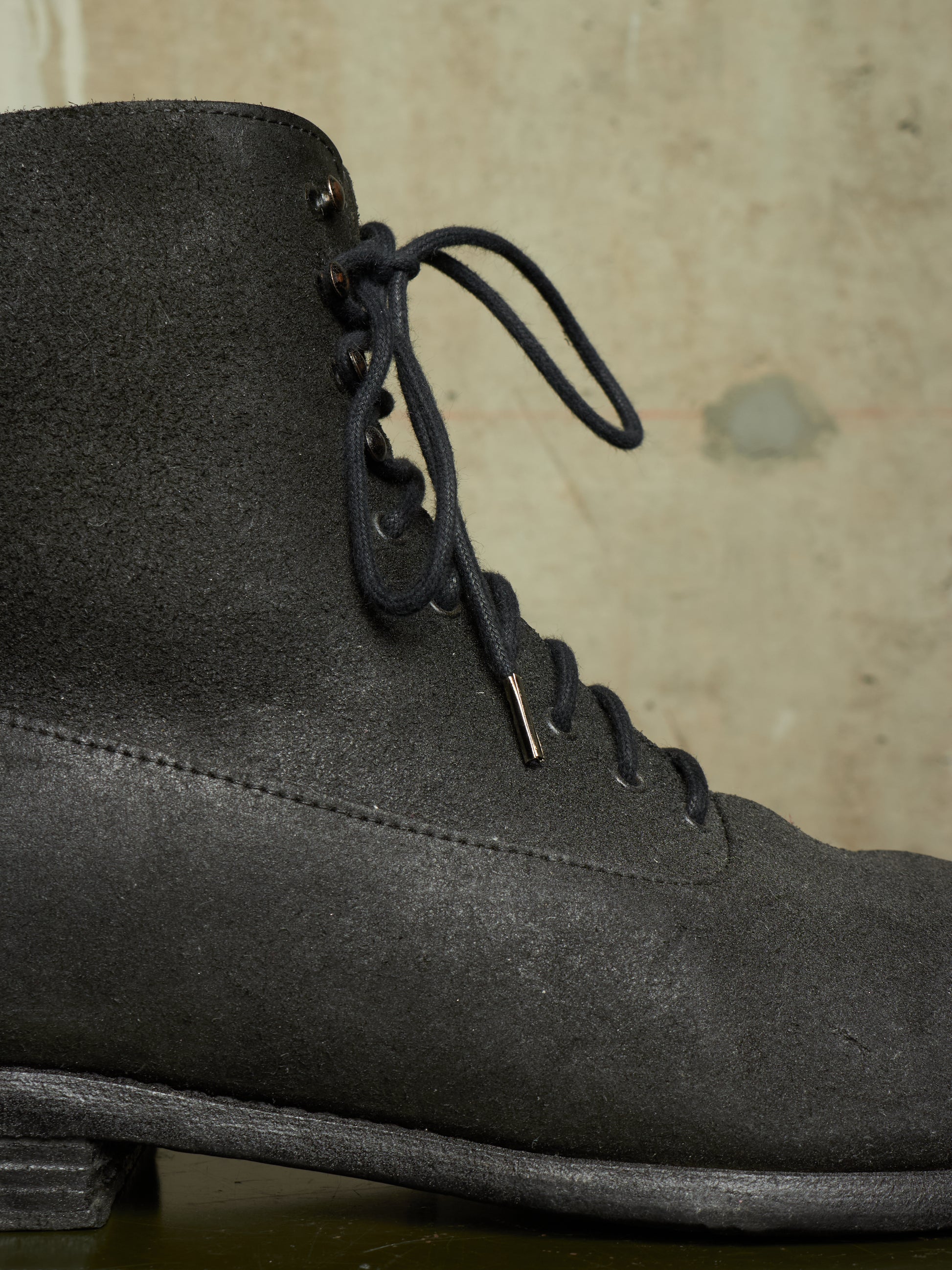 A close-up of a dark gray suede lace-up boot against a textured beige background.