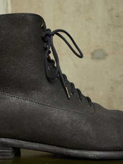 A close-up of a dark gray suede lace-up boot against a textured beige background.
