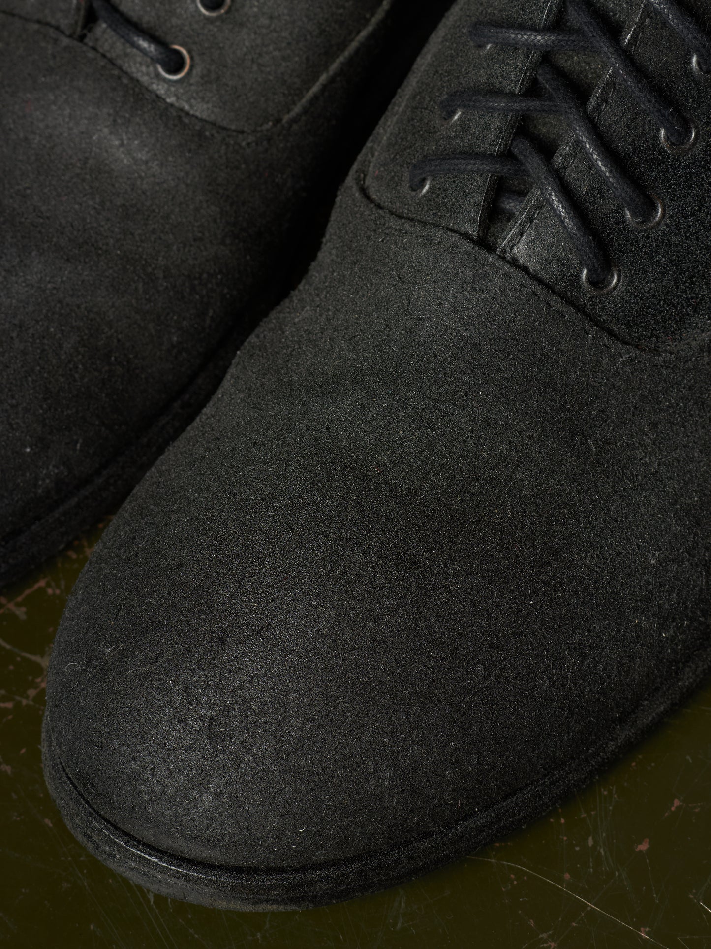 Close-up of a black suede lace-up boot with matching black laces and eyelets, resting on a dark olive-green surface.