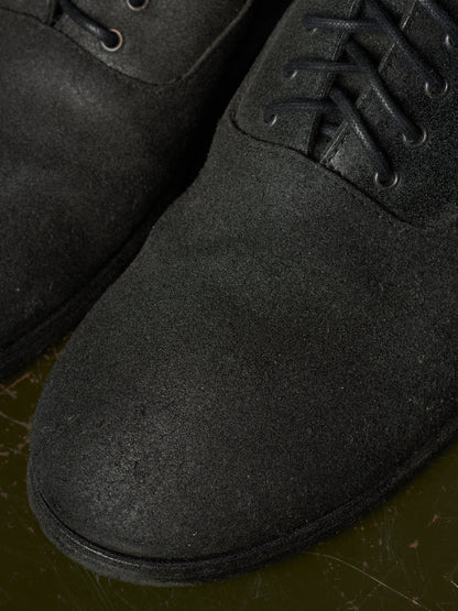 Close-up of a black suede lace-up boot with matching black laces and eyelets, resting on a dark olive-green surface.