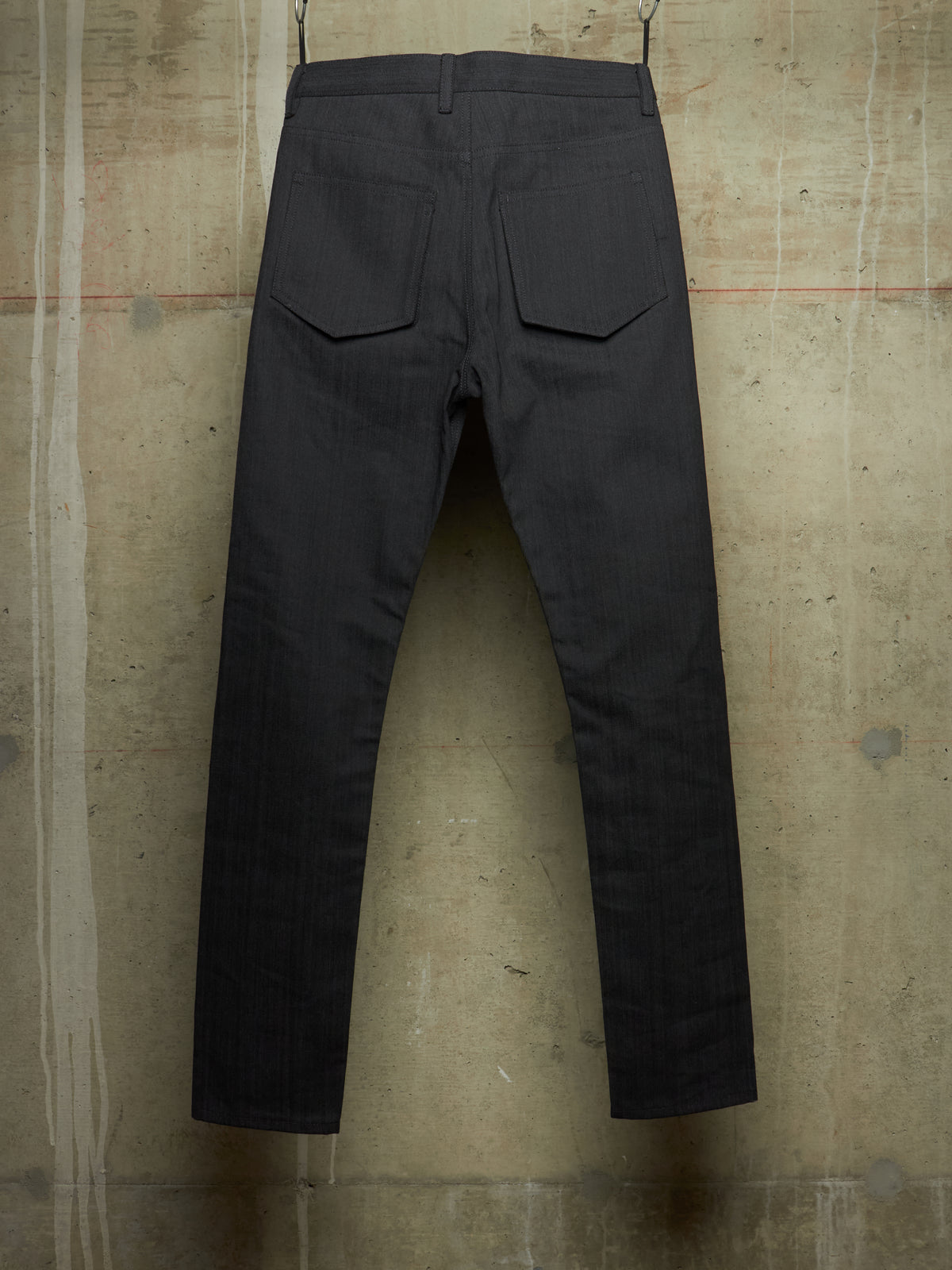 Dark charcoal gray slim-fit jeans shown from the back, hanging against a textured concrete wall.