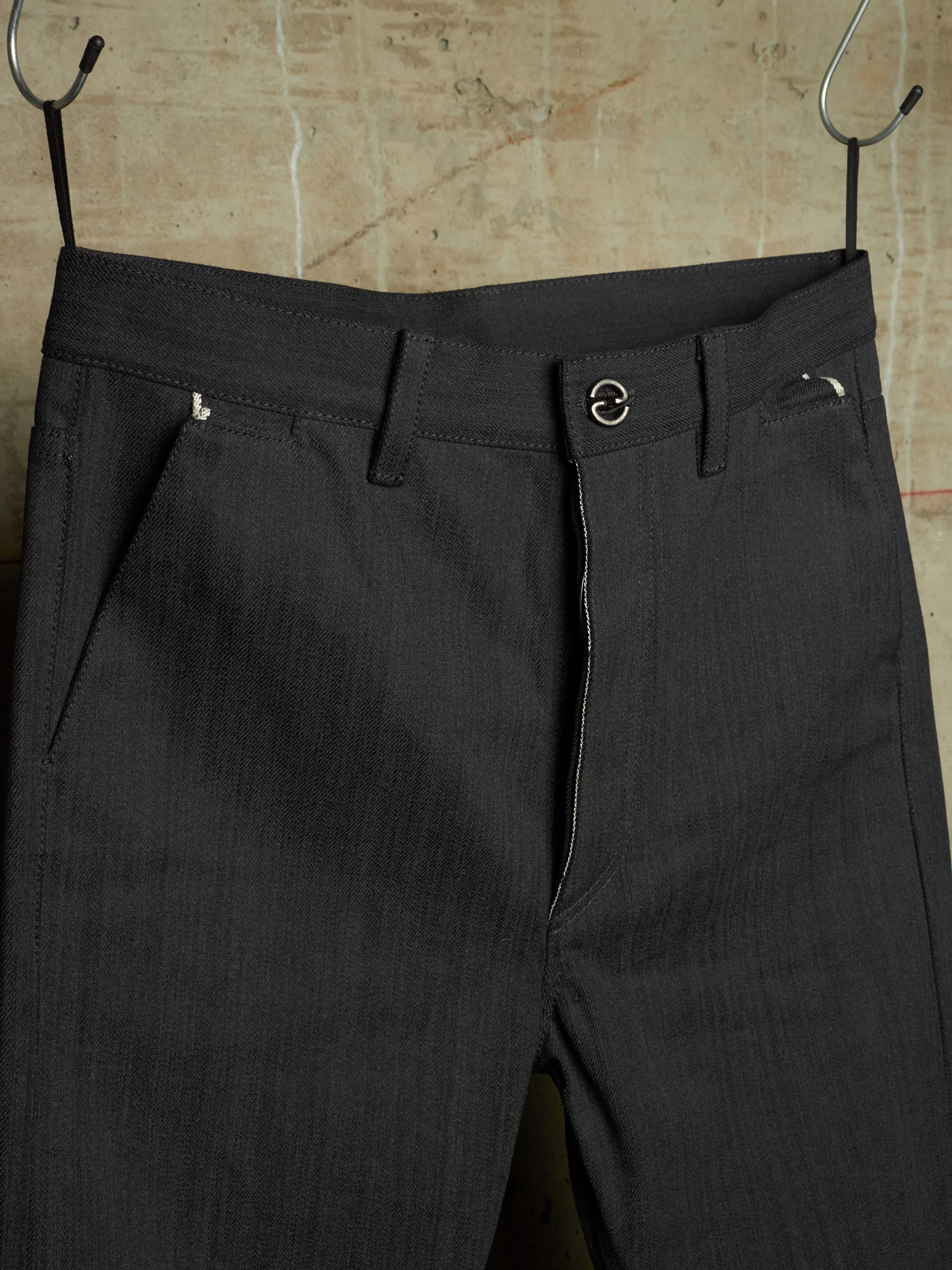 Dark charcoal gray pinstriped trousers hanging from black metal hangers against a textured beige wall.