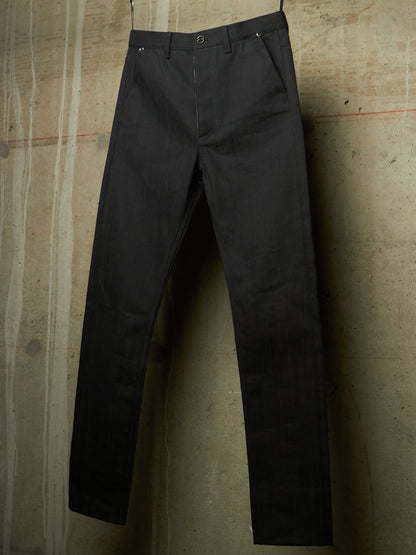 A pair of dark charcoal gray slim-fit trousers hanging against a textured concrete wall.