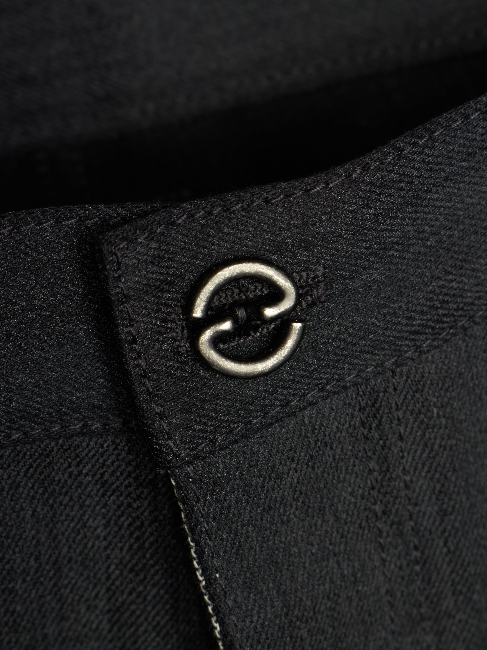 Close-up of a black textured fabric garment featuring a silver metal double-ring hardware detail on a stitched tab.