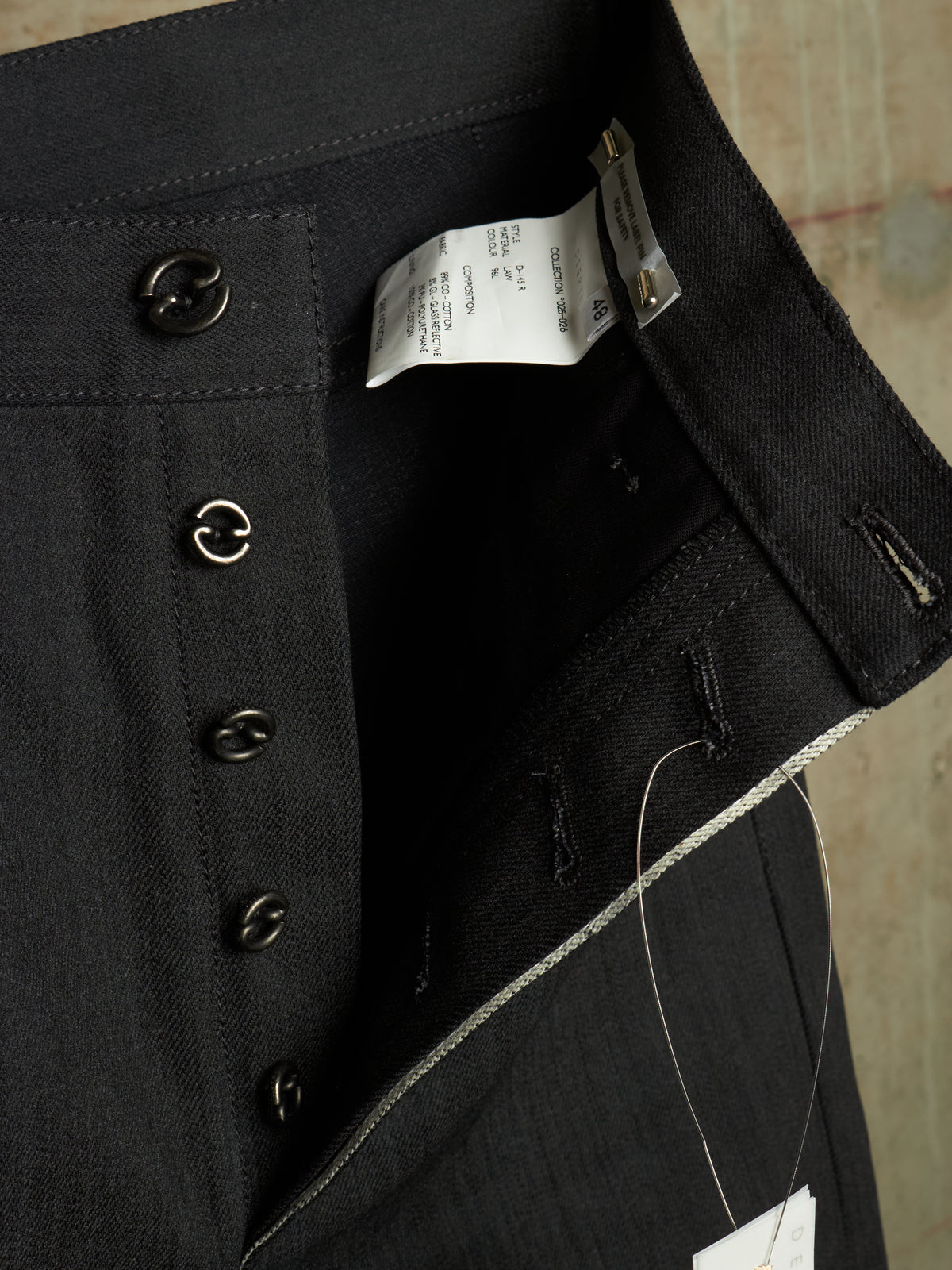 Close-up of a dark charcoal gray or black woven fabric garment, showing the inner waistband with decorative dark buttons, a white size tag, and a thin white thread.
