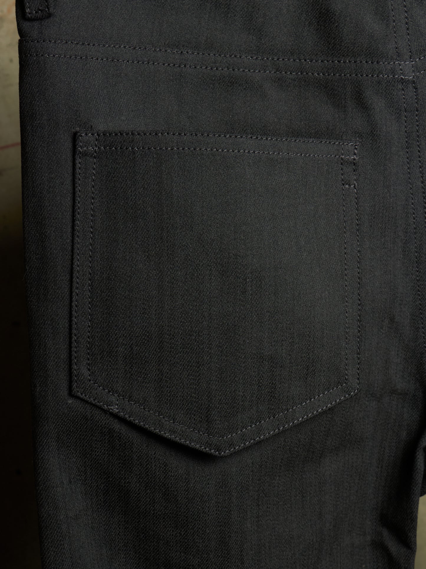 Close-up of the back pocket of dark charcoal gray denim jeans, showing the detailed fabric texture and stitching.