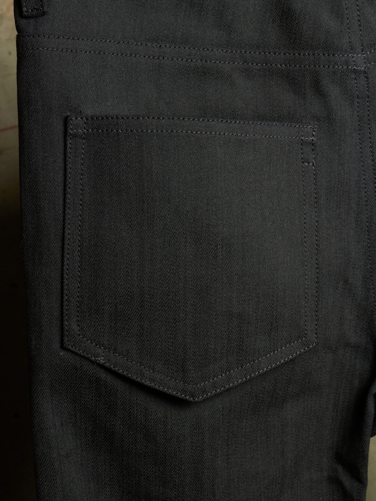 Close-up of the back pocket of dark charcoal gray denim jeans, showing the detailed fabric texture and stitching.