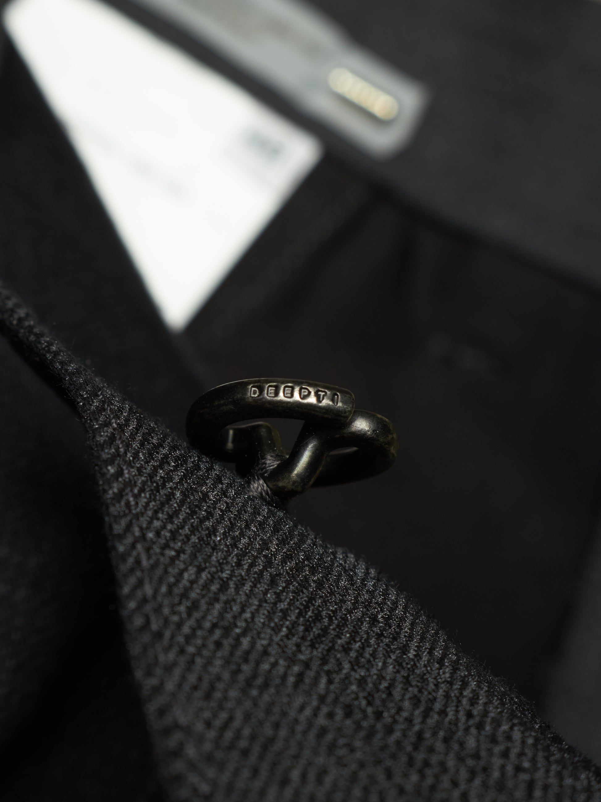 A close-up shot of a black metal ring with engraved 'SEEDS' branding, threaded through a black textured wool or tweed garment, with a blurred black fabric background.