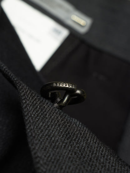 A close-up shot of a black metal ring with engraved 'SEEDS' branding, threaded through a black textured wool or tweed garment, with a blurred black fabric background.