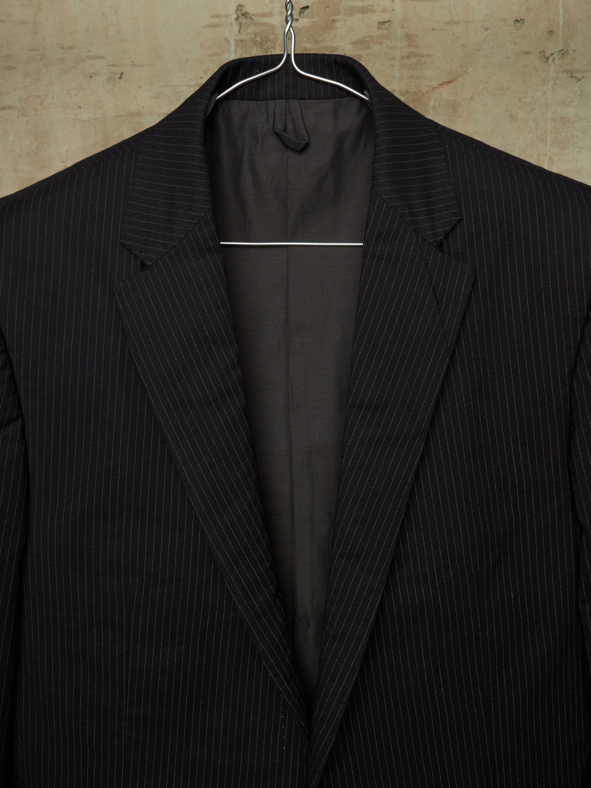 A black pinstripe suit jacket hanging on a metal hanger against a textured beige wall.
