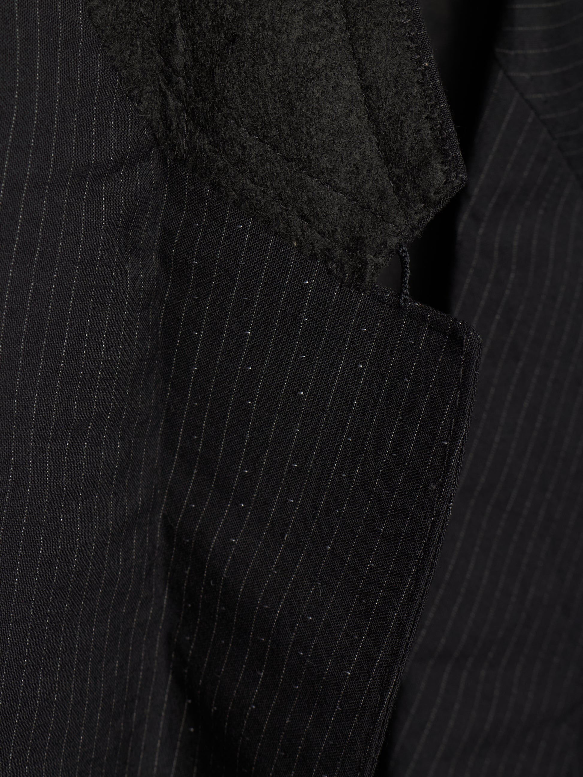 Close-up of a black pinstripe suit jacket showing the textured fabric, fine vertical stripes, and a dark charcoal fuzzy lapel detail.