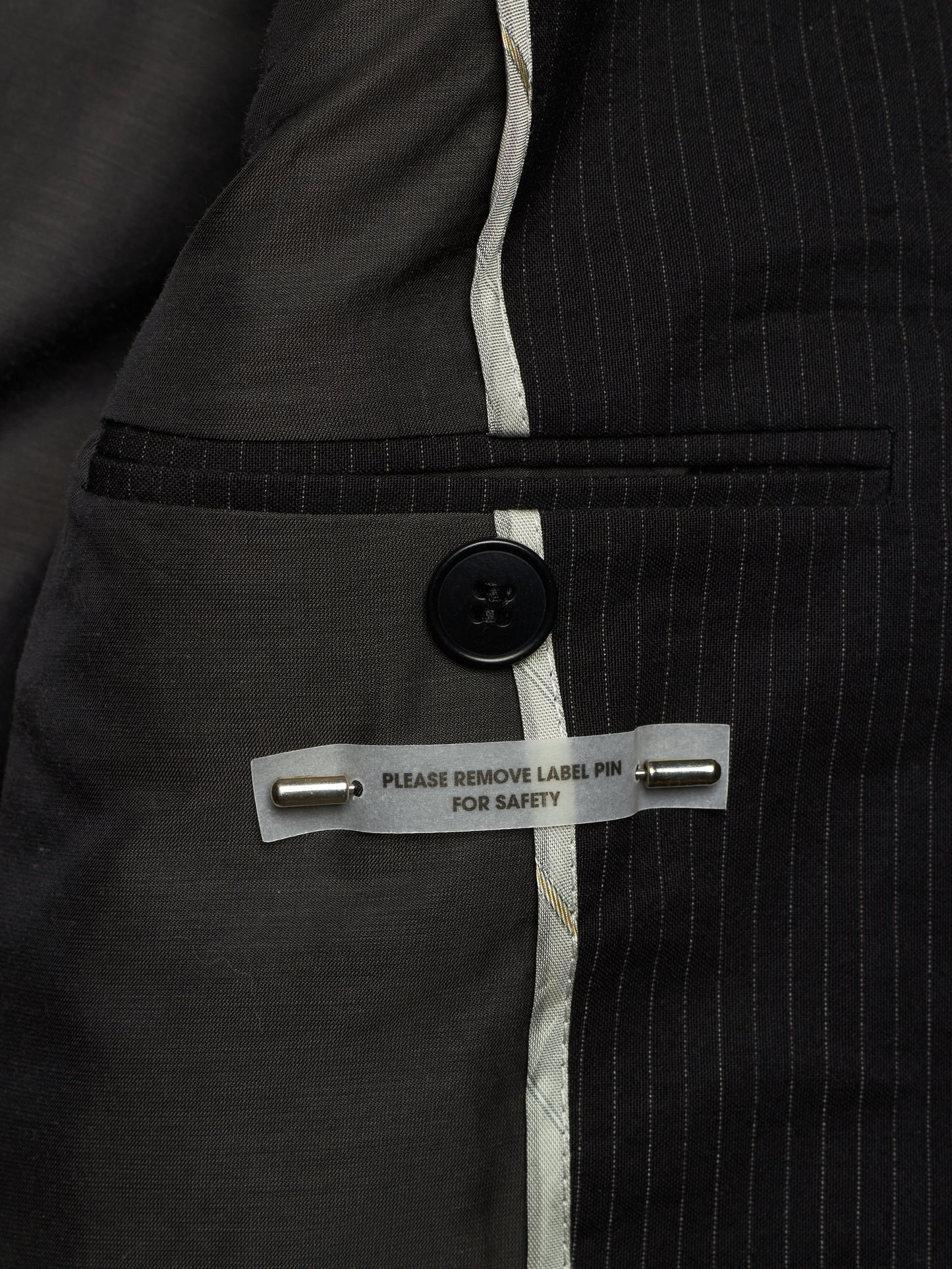 Close-up of a dark gray pinstripe suit jacket showing the interior lining, a buttoned pocket, and a safety pin label.