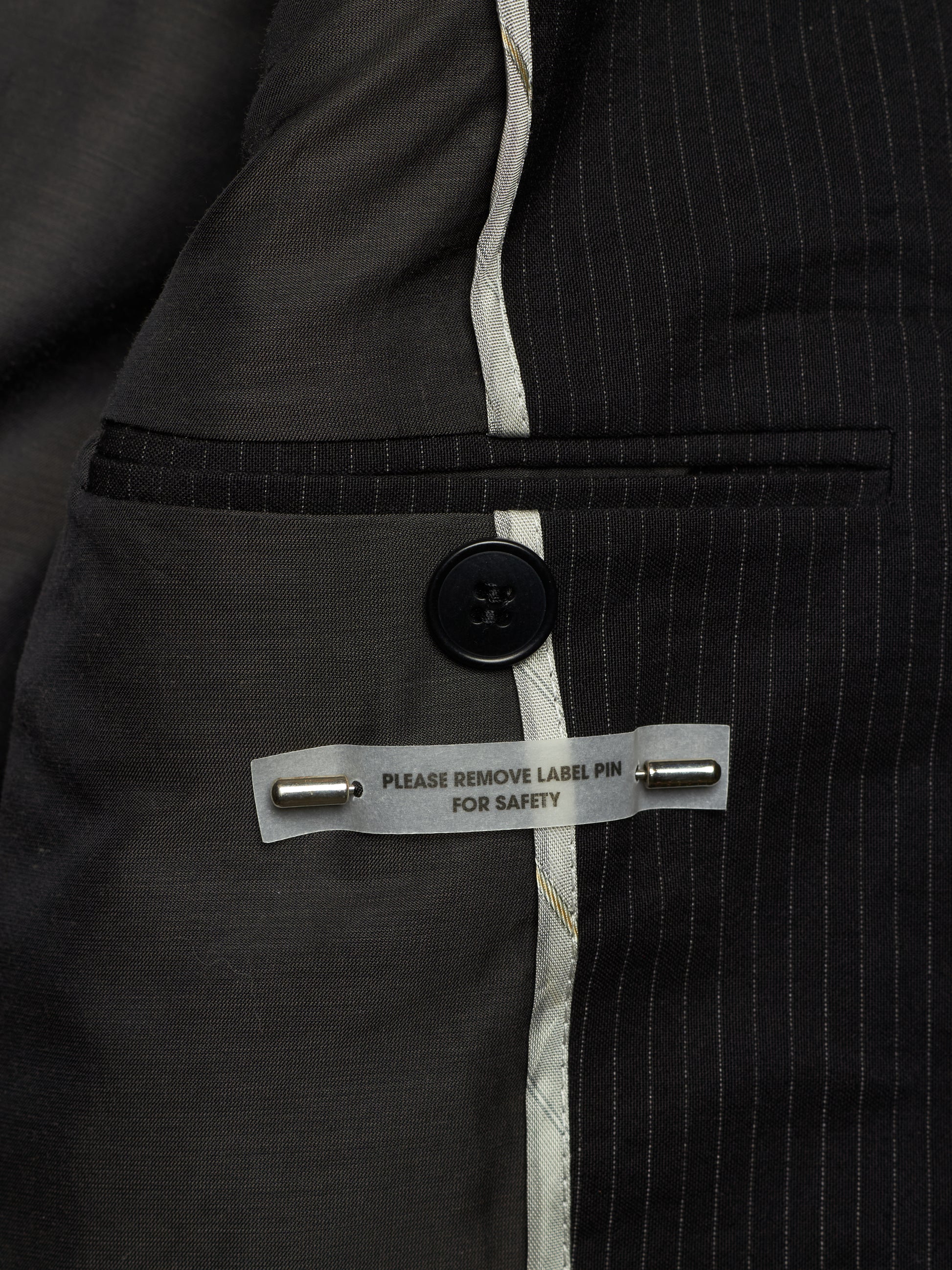 Close-up of a dark gray pinstripe suit jacket showing the interior lining, a buttoned pocket, and a safety pin label.