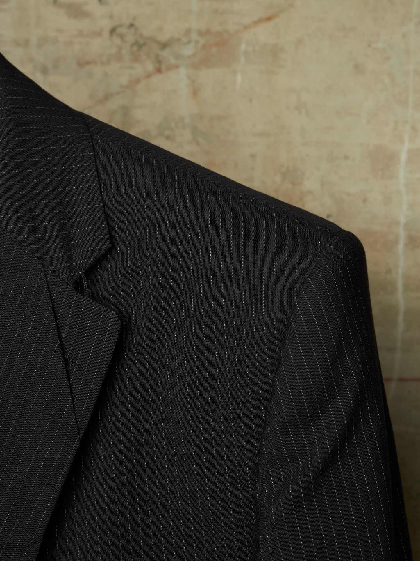 Close-up of a dark charcoal gray pinstripe suit jacket, showing the texture and fine vertical stripes against a textured beige background.