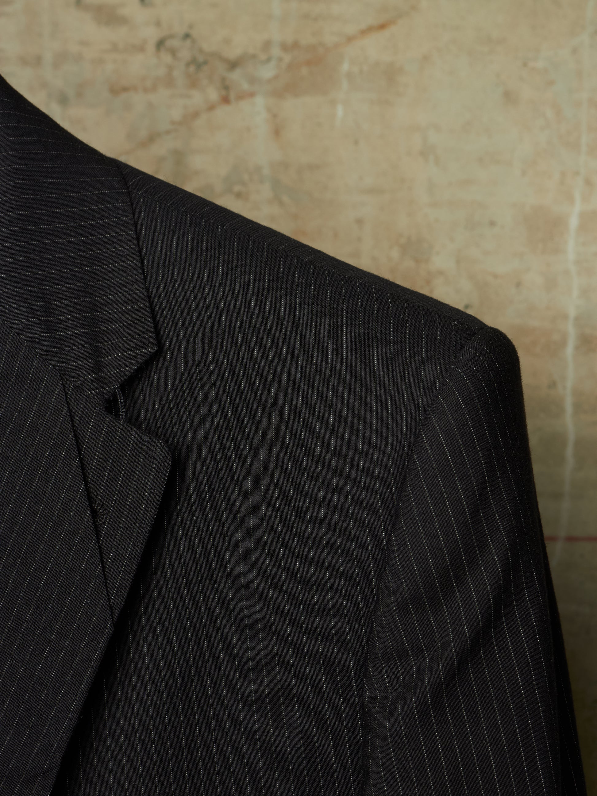Close-up of a dark charcoal gray pinstripe suit jacket, showing the texture and fine vertical stripes against a textured beige background.