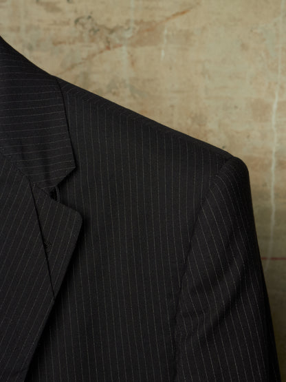 Close-up of a dark charcoal gray pinstripe suit jacket, showing the texture and fine vertical stripes against a textured beige background.