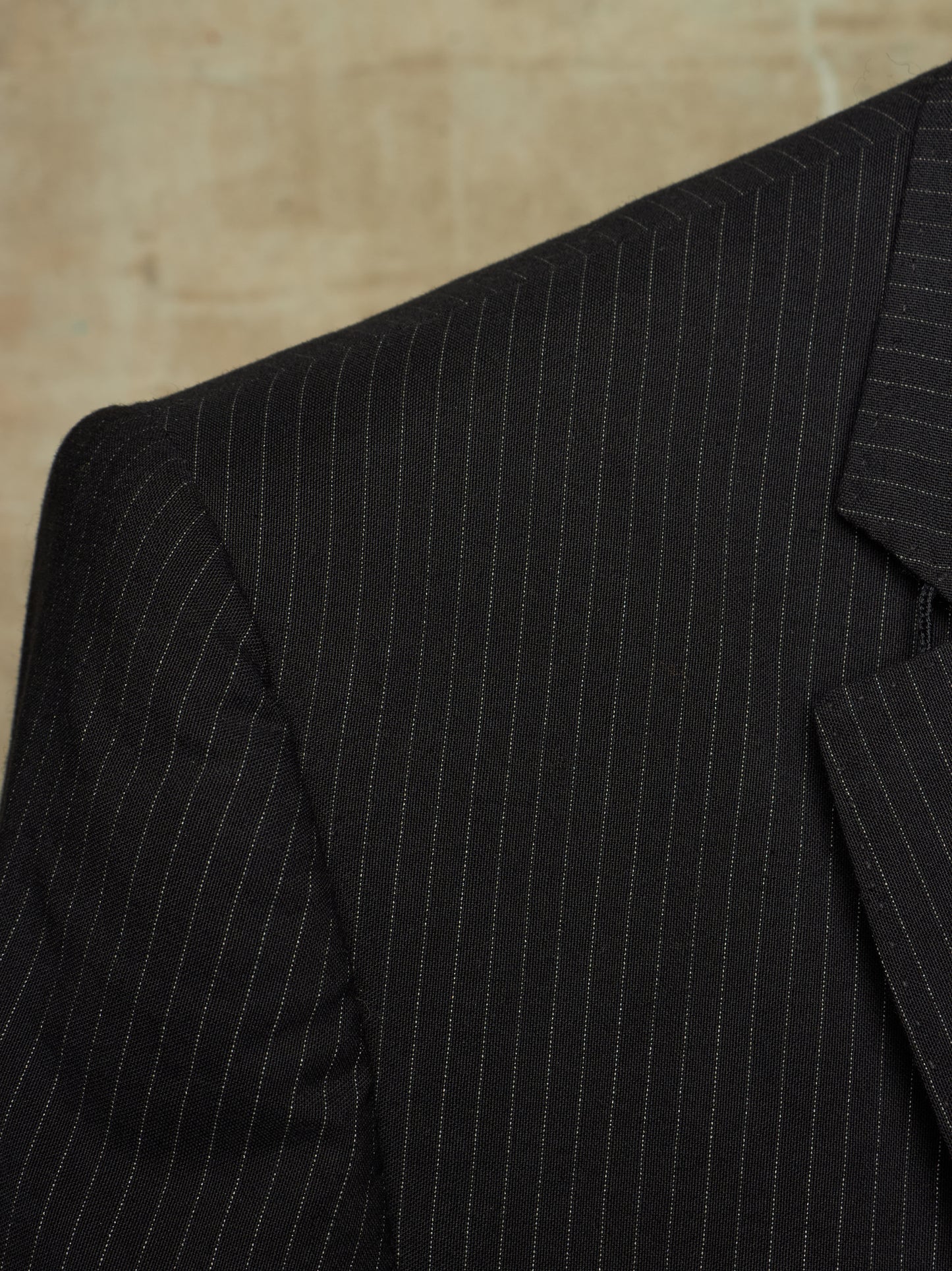 Close-up of a black pinstripe suit jacket showing the fine vertical stripe pattern and lapel detail against a neutral beige background.