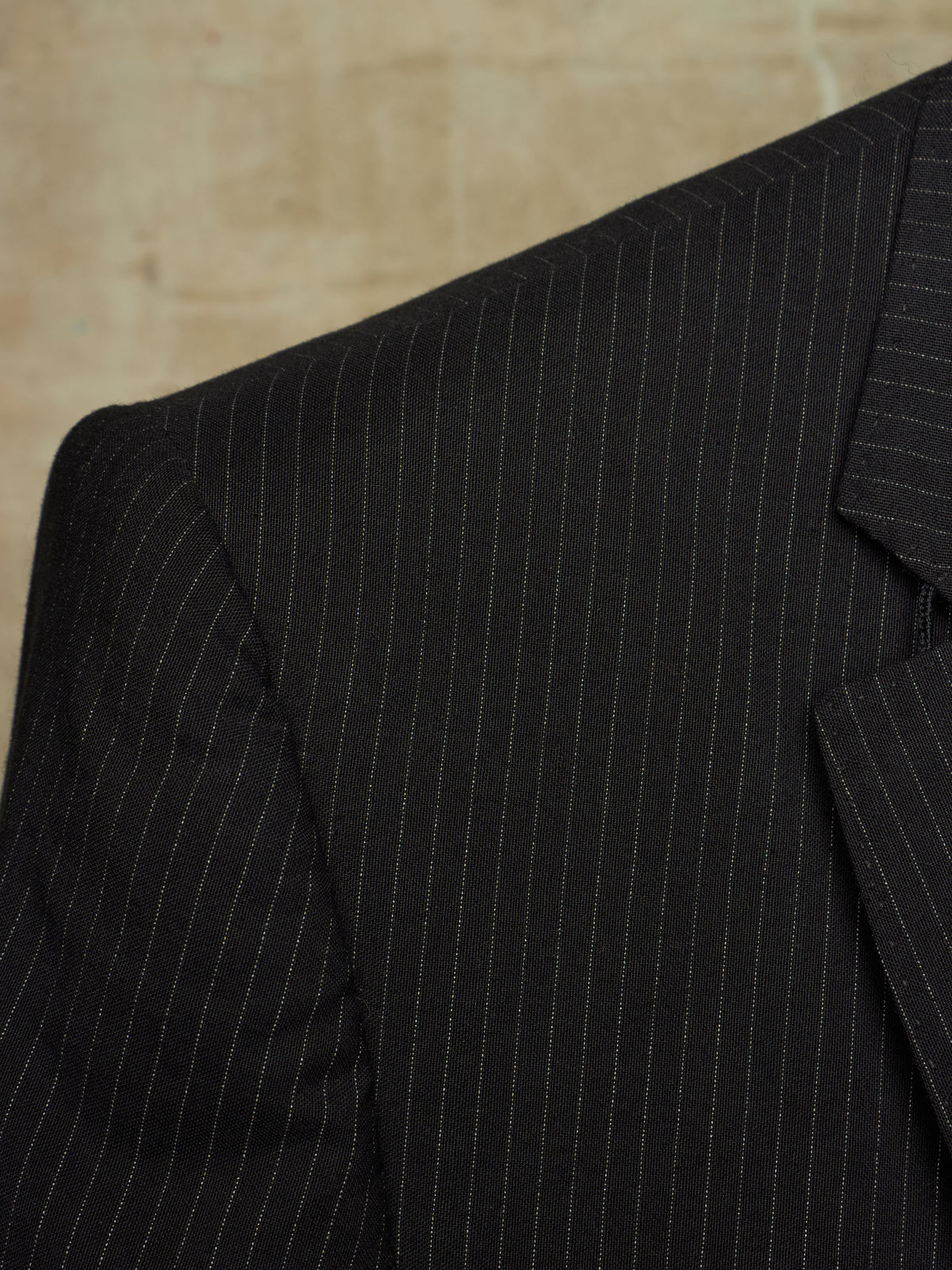 Close-up of a black pinstripe suit jacket showing the fine vertical stripe pattern and lapel detail against a neutral beige background.