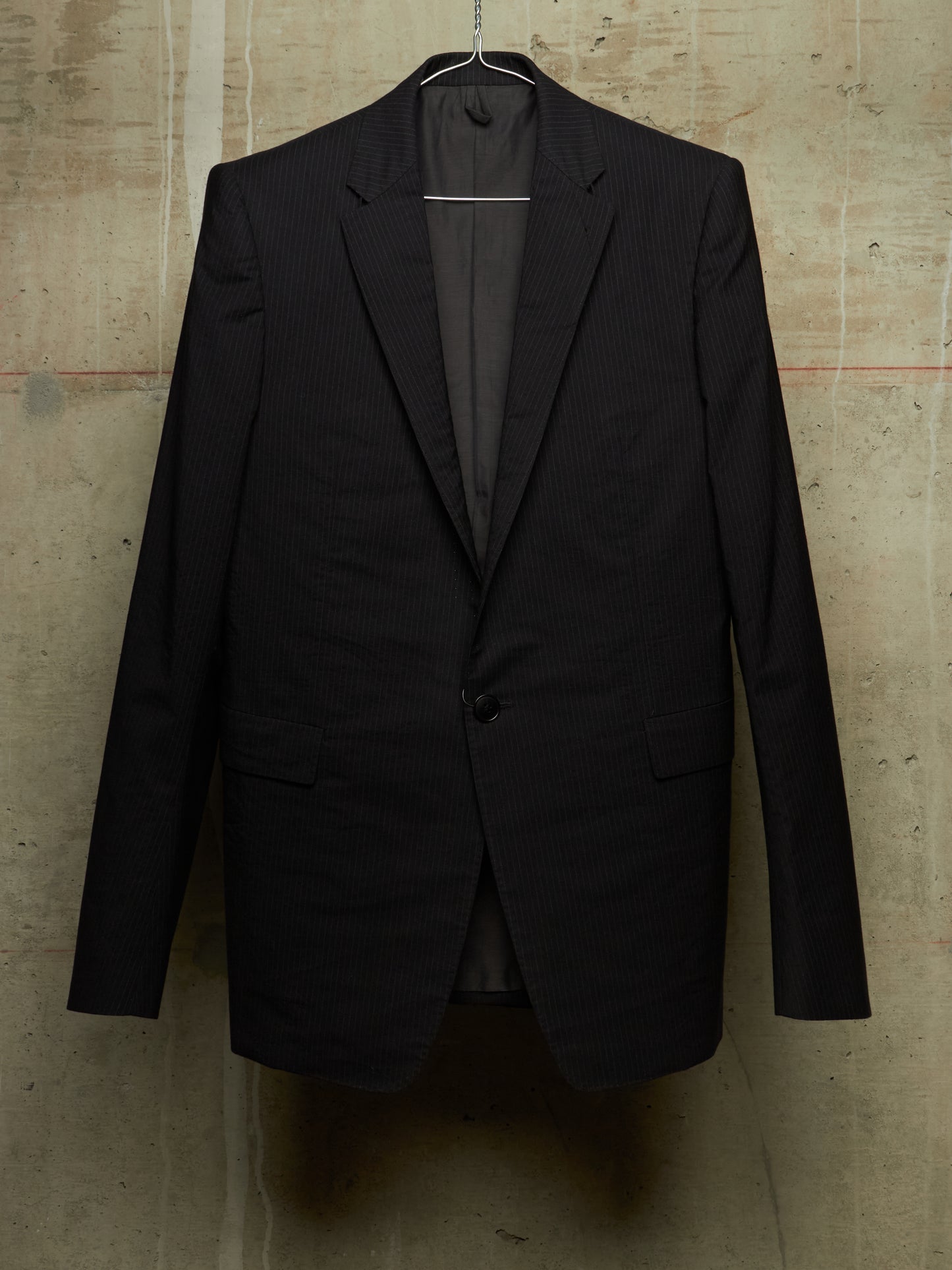 A black pinstripe suit jacket hanging on a metal hanger against a textured concrete wall.